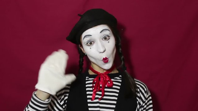 Close-up portrait of a young mime girl. Beautiful mime girl smiles and waves her hand in greeting. Girl in the image of a mime waves her hand when welcoming clients. Portrait on a red background.