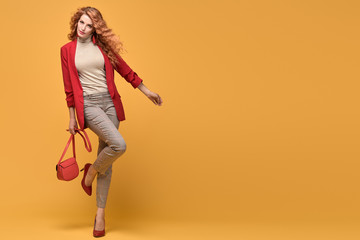 Fashion. Beautiful woman in red jacket, trendy curly hair, make up. Adorable well dressed redhead girl on orange. Gorgeous fashionable lady in red heels, hairstyle, makeup. Creative beauty shot