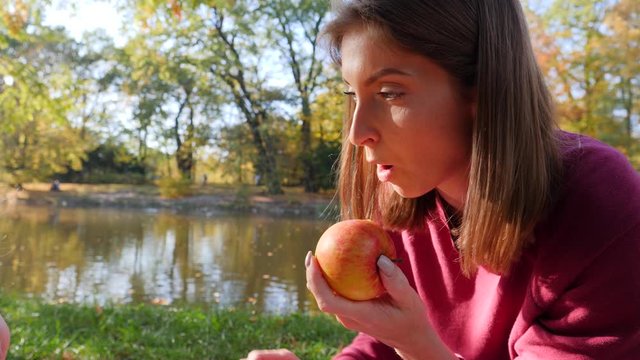 Mother cleans bites a ripe tasty apple fruit gives it to child baby son on rug picnic in a park