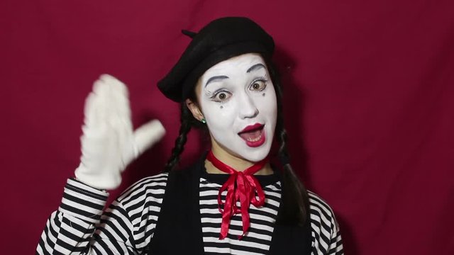 Close-up portrait of a young mime girl. Beautiful mime girl waves her hand, saying goodbye. Girl in the image of a mime waves her hand saying goodbye to clients. Portrait on a red background.
