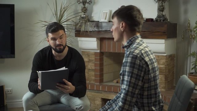 Male Caucasian psychologist in eyeglasses holding notes and talking to his sad patient. Young man undergoing psychological treatment. Individual therapy.