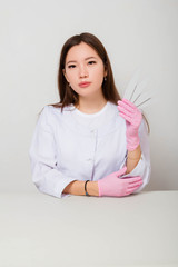 Fototapeta premium Portrait of a girl, master of nails in a white coat and gloves sitting at a table on a white background with nail files in her hands.