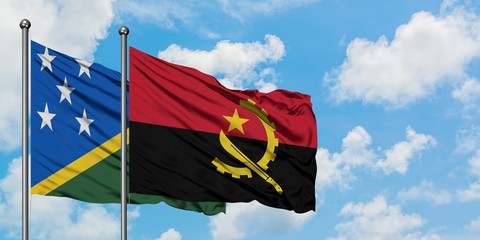 Solomon Islands and Angola flag waving in the wind against white cloudy blue sky together....