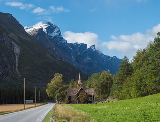 Kors Church, old wooden parish church in Rauma in Romsdal valley, Norway with road E136, green forest and mountain massif Trolltindene, Troll wall Trollveggen. Summer blue sky white clouds.