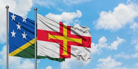 Solomon Islands and Guernsey flag waving in the wind against white cloudy blue sky together....