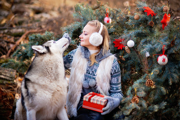 girl sitting in winter park and looking at siberian husky near Christmas tree outdoors.