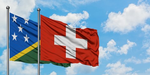 Solomon Islands and Switzerland flag waving in the wind against white cloudy blue sky together. Diplomacy concept, international relations.