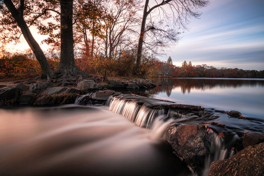 Smooth milky stream of water cascading down rocks from a glassy still lake surrounded by fall foliage. Belmont Lake State Park. 