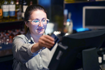 Professional bartender woman in eyeglases registrating new order by cash register. Restaurant worker registrating new order by cash-register. The concept of service. 