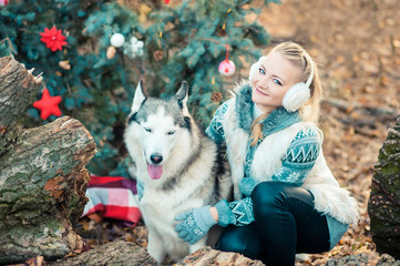 girl sitting in winter park and looking at siberian husky near Christmas tree outdoors.