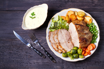 Roasted sliced ham and vegetables: potatoes, carrots, brussel sprouts, cabbage and green peas on black wooden background