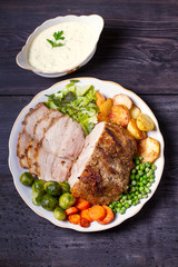 Roasted sliced ham and vegetables: potatoes, carrots, brussel sprouts, cabbage and green peas on black wooden background