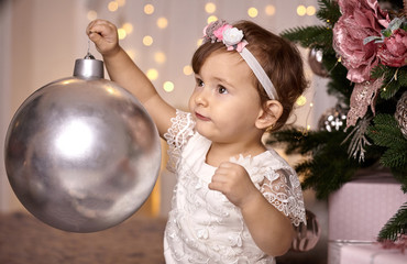 Cute toddler girl playing with a big silver ball to decorate a Christmas tree