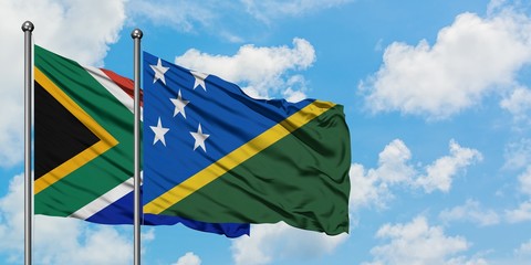 South Africa and Solomon Islands flag waving in the wind against white cloudy blue sky together....