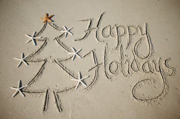 Eco-friendly Happy Holidays message handwritten in smooth sand with simple holiday tree decorated with starfish on the beach