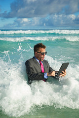 Young businessman in a full suit having a rough time using his tablet computer in crashing waves on the shore of a tropical beach