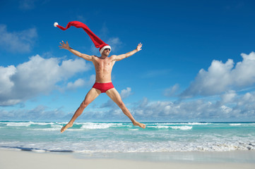 Thin man with extra long Santa hat jumping in red swimming briefs on the shore of a beach in a tropical Christmas celebration