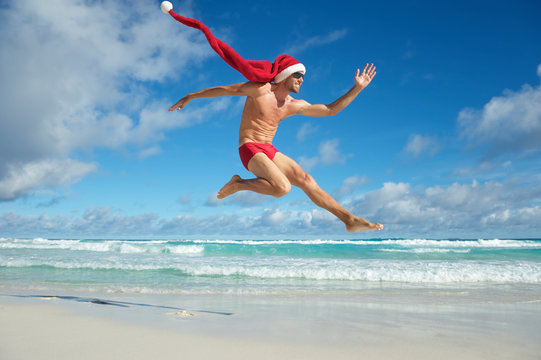Slim man with extra long Santa hat jumping in red swimming briefs on the shore of a beach in a tropical Christmas celebration