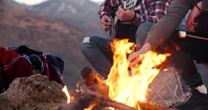 Four multiethnic students sitting near campfire on top of mountain, roasting marshmallows, dancing and listening to guitar music, expressing positive emotions - friendship, love concept 4k footage