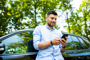 Checking social media. Young attractive smiling man with beard standing near his car and holding...