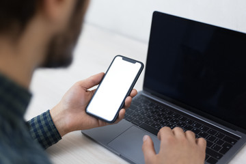 Close-up of male hand, using laptop and holding smartphone with mockup.