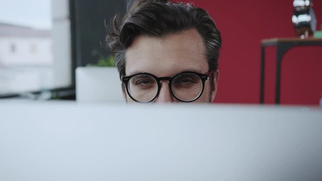 Fun scene in the office. Close-up tricky manager director appearing behind monitor winking with eye flirting on camera while working.