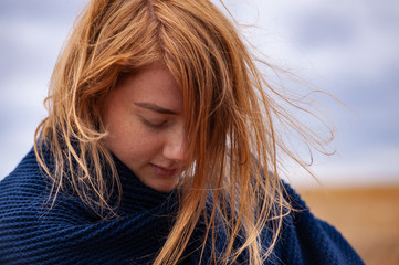 young girl with red hair and sun kissed face posing on the yellow autumn field under dark cloudy sky wrapped in cosy dark blue plaid