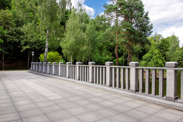 view of a bridge with stone tiles and granite railings with square columns on background a park with green trees and sky, nobody.