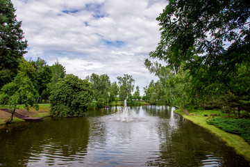 fountain in the pond on the park with green plants and blue sky with clouds.