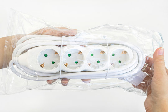 Woman hand holding new four outlets power extension cord in a transparent plastic pakage over white table. Electric power strip for home or office. Household appliances.