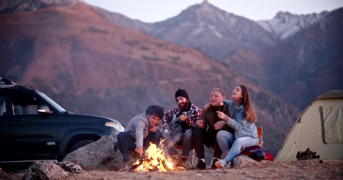 Group of four hipster frends relaxing on top of a mountain, sitting near bonfire, roasting marshmallows and enjoying their time - friendship, youth, love concept 4k footage