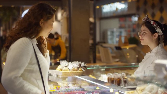 Young beautiful woman walking to sweets corner in food mall, looking at assortment in showcase and discussing cakes with cheerful saleswoman