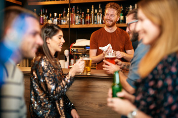 Handsome caucasian barman staning in bar and wiping pint and talking to customers. Customers drinking beer. Nightlife.