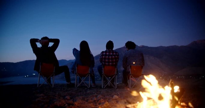 Silhouettes of four friends sitting in chairs near bonfire on top of mountains, watching the sun set and chatting, enjoying their trip - friendship, youth concept 4k footage