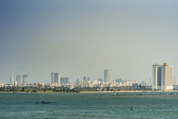 Da Nang, Vietnam - March 10, 2019: Tien Sa Port in Da Nang Bay. City skyline as urban stone band separating azure sea from light blue sky. Some green foliage along yellow beach line. small fishing ves