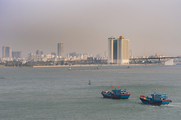 Da Nang, Vietnam - March 10, 2019: Tien Sa Port in Da Nang Bay. 2 blue and red fishing vessels return home in evening. Cityscape and long on ramp of Thuan Phuoc bridge on horizon.