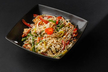 Chicken noodles bowl with vegetable. Udon stir-fry noodles with chicken meat and sesame in bowl on dark stone background copy space. Spaghetti pasta with chicken and vegetable. Horizontal photo.
