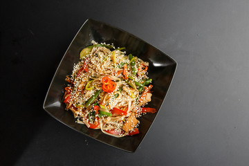 Chicken noodles bowl with vegetable. Udon stir-fry noodles with chicken meat and sesame in bowl on dark stone background copy space. Spaghetti pasta with chicken and vegetable. Horizontal photo.