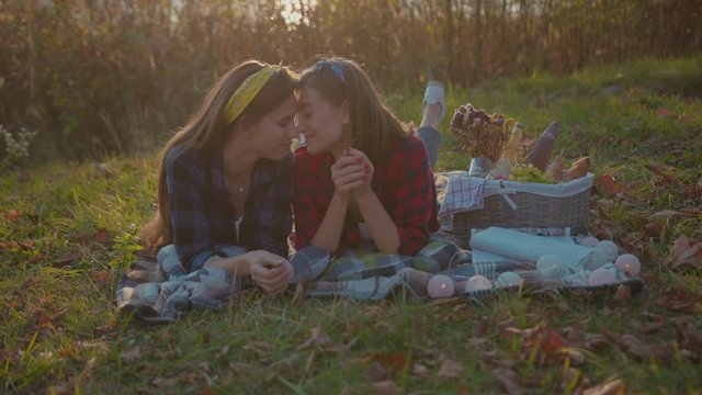 Adorable loving couple of girls lying on grass hugging tight together at sunset. Woman eating fruits cuddling with her sweetheart in beautiful evening scenery.