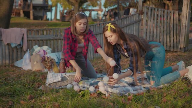 Attractive young women making picnic decoration with lights blankets and fruits staying together in the countryside spending weekend evening.