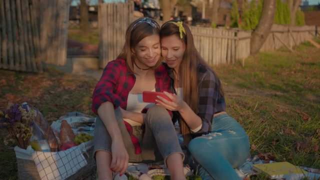 Beautiful cute young women using smartphone sitting on blanket grass in the countryside. Couple of girls browsing internet content mobile phone enjoying picnic time in autumn.