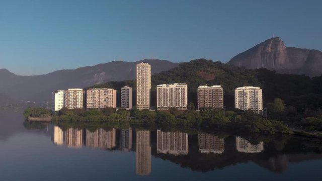 Slow aerial backing off of high rise residential apartment buildings reflected in the city lake Lagoa Rodrigo de Freitas in Rio de Janeiro with the Corcovado mountain in the background at sunrise