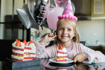 Indoor shot of pretty joyful little girl with blonde hair blowing out the candle, celebrate 6 years...