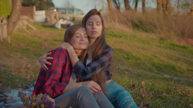 Pleased attractive woman hugging her dear sweetheart girl relaxing together on grass at beautiful sunset in the countryside. Romance and dating concept.
