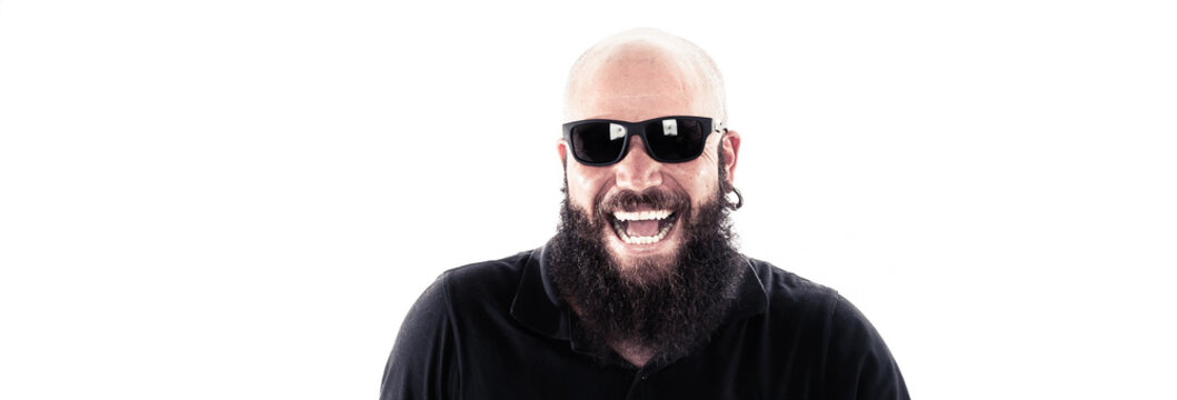 Um homem careca e de barba longa que est&aacute; usando um &oacute;culos escuro e brincos de argola dando risada ou gargalhadas como um louco em foto com fundo branco de est&uacute;dio. Adulto, Hipster, Roqueiro.