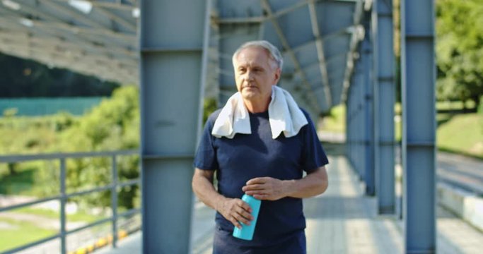 Old Caucasian grey-haired man walking at the stadium after jogging and drinking water while resting and smling. Outdoor.