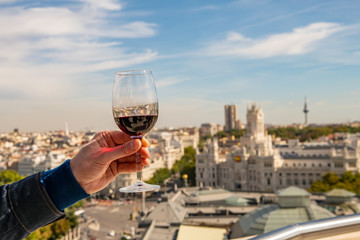 A glass of red wine held up for a toast on a rooftop overlooking the beautiful cityscape of Madrid,...