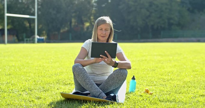 Caucasian senior woman sitting on the mat on the freen grass of the stadium and using tablet device, tapping, scrolling and watching something on the screen about sport in the morning.