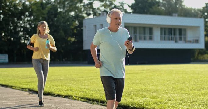 Caucasian senior sporty msn and woman running on a sunny summer day and listening to the music while keeping themselves in good shapes.