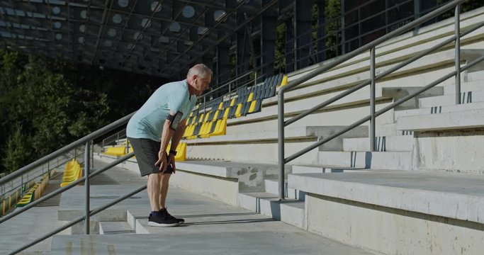Old Caucasian man at retirement stepping up the steps at the stadium and stops to have a rest, then keeps on going. Outdoors.